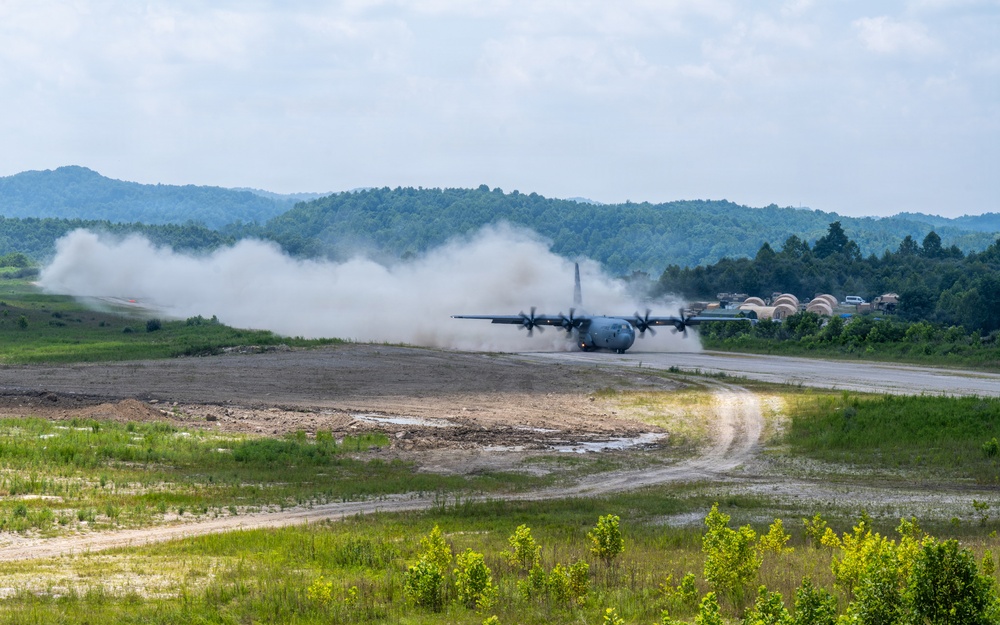 A military turboprop transport aircraft, consistent with a C-130 Hercules, is landing or taking off on a short unpaved or semi-prepared airstrip, generating significant dust and smoke clouds. The setting appears to be a forward operating base or austere airfield surrounded by forested hills and green vegetation. Several dome-shaped military shelters or hangars are visible in the background to the right.