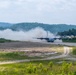 A military turboprop transport aircraft, consistent with a C-130 Hercules, is landing or taking off on a short unpaved or semi-prepared airstrip, generating significant dust and smoke clouds. The setting appears to be a forward operating base or austere airfield surrounded by forested hills and green vegetation. Several dome-shaped military shelters or hangars are visible in the background to the right.