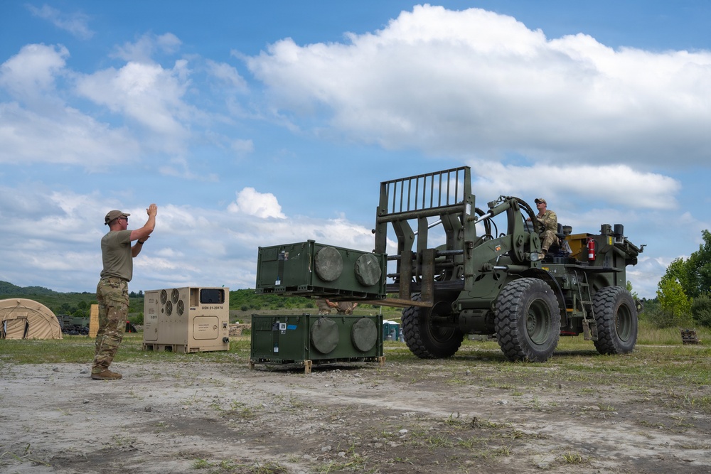 Military personnel are conducting a logistics or equipment-handling operation at an outdoor field site. A soldier on the left is using hand signals to guide a forklift operator who is maneuvering military equipment containers. The setting appears to be a forward operating base or training area with tents and vehicles visible in the background.