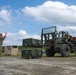 Military personnel are conducting a logistics or equipment-handling operation at an outdoor field site. A soldier on the left is using hand signals to guide a forklift operator who is maneuvering military equipment containers. The setting appears to be a forward operating base or training area with tents and vehicles visible in the background.