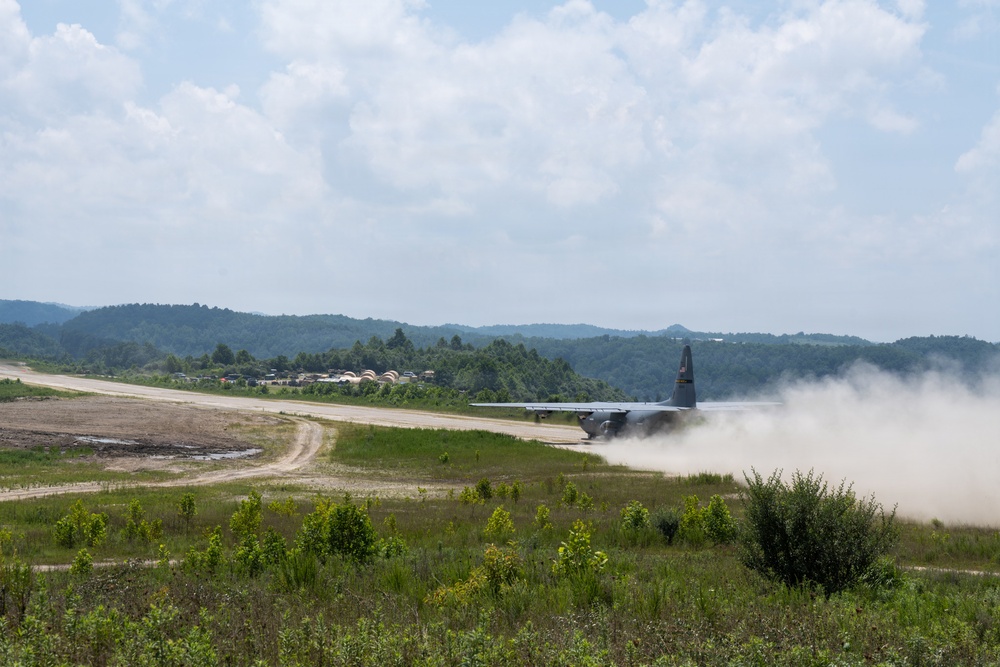 A military transport aircraft, appearing to be a C-130 Hercules, is landing or taking off on an unpaved dirt/gravel airstrip, generating a large dust cloud behind it. The airstrip is situated in a rural, hilly area surrounded by dense green vegetation and forested hills. Several military structures or hangars are visible in the background near the runway.