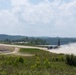 A military transport aircraft, appearing to be a C-130 Hercules, is landing or taking off on an unpaved dirt/gravel airstrip, generating a large dust cloud behind it. The airstrip is situated in a rural, hilly area surrounded by dense green vegetation and forested hills. Several military structures or hangars are visible in the background near the runway.