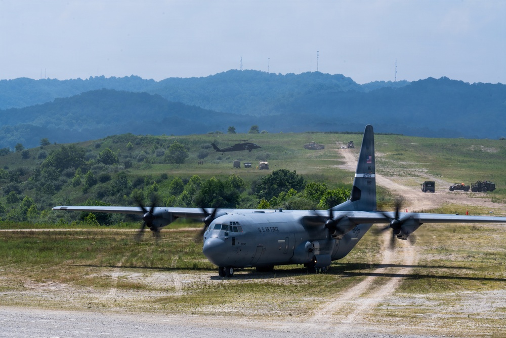 A U.S. Air Force C-130J Super Hercules tactical transport aircraft is operating on an unpaved forward airstrip in a hilly, vegetated terrain. Multiple military ground vehicles are positioned along a dirt road in the background, and a military helicopter (likely a UH-60 Black Hawk) is visible in flight. The scene depicts a joint military exercise or tactical airlift operation.