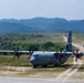 A U.S. Air Force C-130J Super Hercules tactical transport aircraft is operating on an unpaved forward airstrip in a hilly, vegetated terrain. Multiple military ground vehicles are positioned along a dirt road in the background, and a military helicopter (likely a UH-60 Black Hawk) is visible in flight. The scene depicts a joint military exercise or tactical airlift operation.