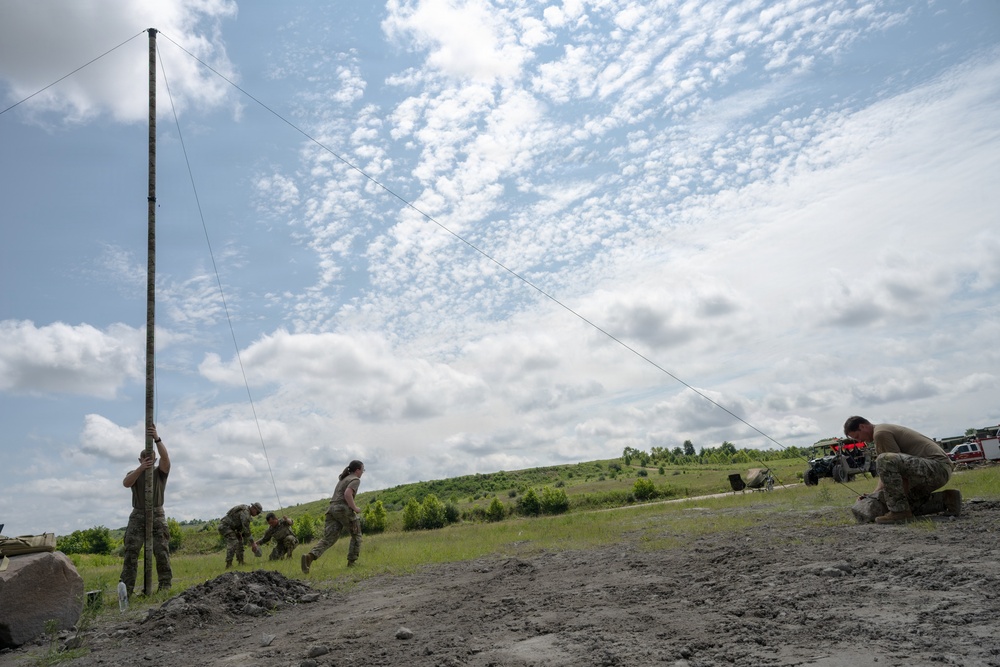 A group of military personnel in camouflage uniforms are erecting a tall antenna or mast pole in an open field area with disturbed soil. Guy-wires are being tensioned and staked to stabilize the structure. The setting appears to be a field training exercise or communications setup operation.