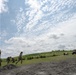 A group of military personnel in camouflage uniforms are erecting a tall antenna or mast pole in an open field area with disturbed soil. Guy-wires are being tensioned and staked to stabilize the structure. The setting appears to be a field training exercise or communications setup operation.