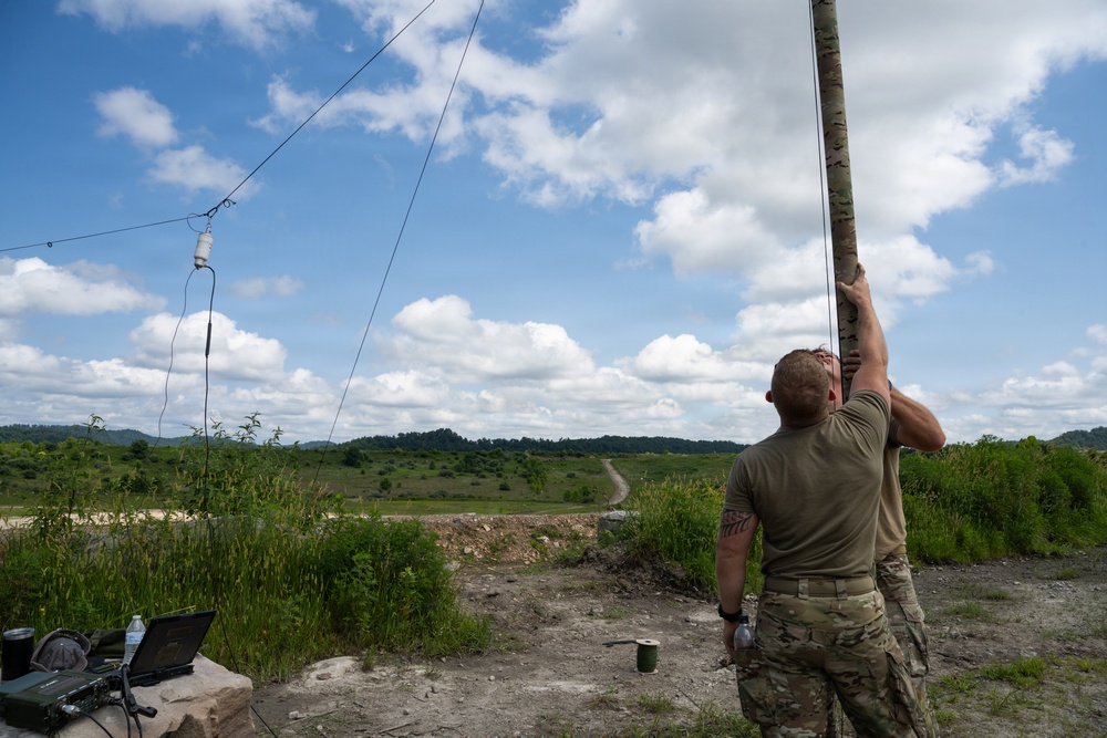 A soldier in military camouflage (MultiCam/OCP pattern) is erecting a tall antenna mast in an outdoor field environment. A wire antenna is already strung and suspended via an insulator, connected to the mast. Communications equipment including a laptop and radio gear is visible on the ground nearby.