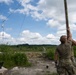 A soldier in military camouflage (MultiCam/OCP pattern) is erecting a tall antenna mast in an outdoor field environment. A wire antenna is already strung and suspended via an insulator, connected to the mast. Communications equipment including a laptop and radio gear is visible on the ground nearby.