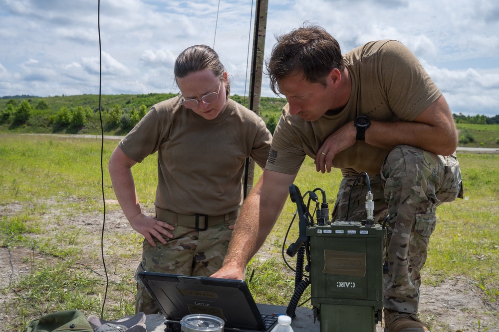 Two military personnel in OCP (Operational Camouflage Pattern) uniforms are working together at a field station, examining a ruggedized Getac laptop. They are operating alongside military communications equipment, including what appears to be a tactical radio unit labeled 'CARC', with an antenna mast visible in the background. The setting is an open outdoor training or operational area with grassland and treeline visible.