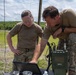 Two military personnel in OCP (Operational Camouflage Pattern) uniforms are working together at a field station, examining a ruggedized Getac laptop. They are operating alongside military communications equipment, including what appears to be a tactical radio unit labeled 'CARC', with an antenna mast visible in the background. The setting is an open outdoor training or operational area with grassland and treeline visible.