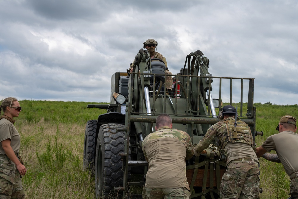 Military personnel in multicam/OCP uniforms are working together to maneuver a piece of heavy military equipment, likely an artillery piece or towed vehicle, in an open grassy field. One soldier is seated at the operator position on top of the equipment while several others push or guide it from behind and the side. The setting appears to be a training exercise conducted in a rural outdoor environment under overcast skies.