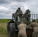 Military personnel in multicam/OCP uniforms are working together to maneuver a piece of heavy military equipment, likely an artillery piece or towed vehicle, in an open grassy field. One soldier is seated at the operator position on top of the equipment while several others push or guide it from behind and the side. The setting appears to be a training exercise conducted in a rural outdoor environment under overcast skies.