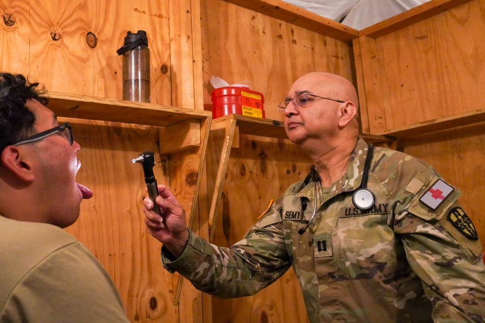 A U.S. Army medical officer, identified by name tape as 'SEMY' and wearing Captain rank insignia, is conducting a medical examination of a patient in what appears to be a field medical facility. The physician is holding an otoscope/ophthalmoscope and examining the patient's throat or mouth area. The setting is a makeshift wooden structure consistent with a forward operating base or field clinic.