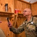 A U.S. Army medical officer, identified by name tape as 'SEMY' and wearing Captain rank insignia, is conducting a medical examination of a patient in what appears to be a field medical facility. The physician is holding an otoscope/ophthalmoscope and examining the patient's throat or mouth area. The setting is a makeshift wooden structure consistent with a forward operating base or field clinic.