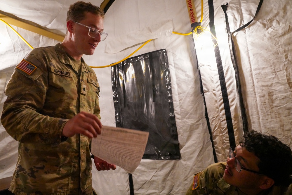 Two U.S. Army soldiers are inside a military field tent, with one standing soldier named 'Bernabei' holding and reviewing a document while speaking to a seated soldier. Both wear OCP (Operational Camouflage Pattern) uniforms with U.S. flag patches. The setting appears to be a temporary field operations or medical tent with yellow power cables and black structural straps visible.