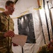 Two U.S. Army soldiers are inside a military field tent, with one standing soldier named 'Bernabei' holding and reviewing a document while speaking to a seated soldier. Both wear OCP (Operational Camouflage Pattern) uniforms with U.S. flag patches. The setting appears to be a temporary field operations or medical tent with yellow power cables and black structural straps visible.
