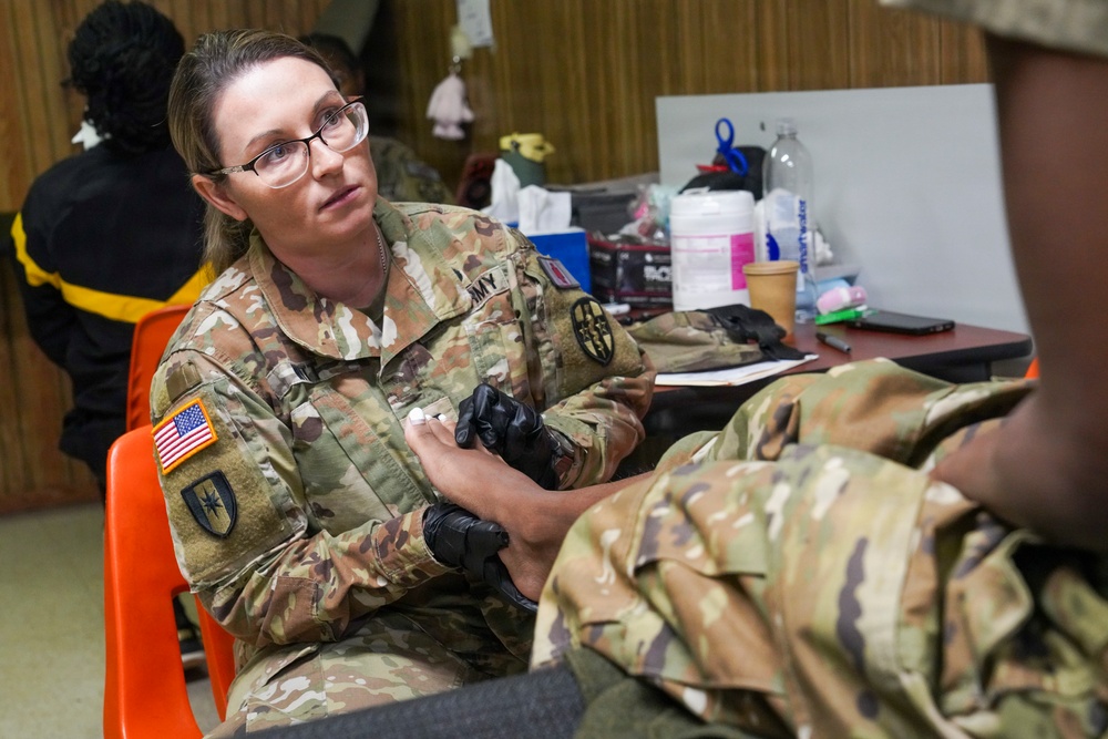 A female U.S. Army soldier wearing OCP (Operational Camouflage Pattern) uniform and black medical gloves is providing medical care or treatment to another soldier's foot or lower leg. She is wearing glasses and appears focused on her task in what looks like a makeshift medical station or community facility. Medical supplies and equipment are visible on a table in the background.