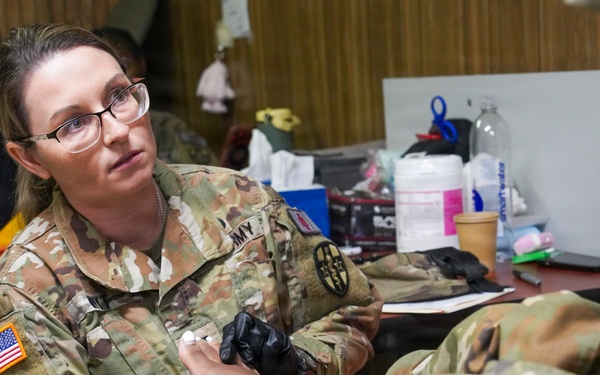 A female U.S. Army soldier wearing OCP (Operational Camouflage Pattern) uniform and black medical gloves is providing medical care or treatment to another soldier's foot or lower leg. She is wearing glasses and appears focused on her task in what looks like a makeshift medical station or community facility. Medical supplies and equipment are visible on a table in the background.