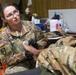 A female U.S. Army soldier wearing OCP (Operational Camouflage Pattern) uniform and black medical gloves is providing medical care or treatment to another soldier's foot or lower leg. She is wearing glasses and appears focused on her task in what looks like a makeshift medical station or community facility. Medical supplies and equipment are visible on a table in the background.