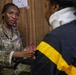 A U.S. Army soldier, identified by name tape as 'KONNEH' and wearing an OCP (Operational Camouflage Pattern) uniform with a U.S. flag patch, is engaged in what appears to be a consultation or interview session with a civilian. The soldier is holding a pink pen and appears to be taking notes while speaking with the individual seated across from her. A handwritten note on the wall behind reads 'Laugh Love - CPT KONNEH', suggesting this is her workspace.
