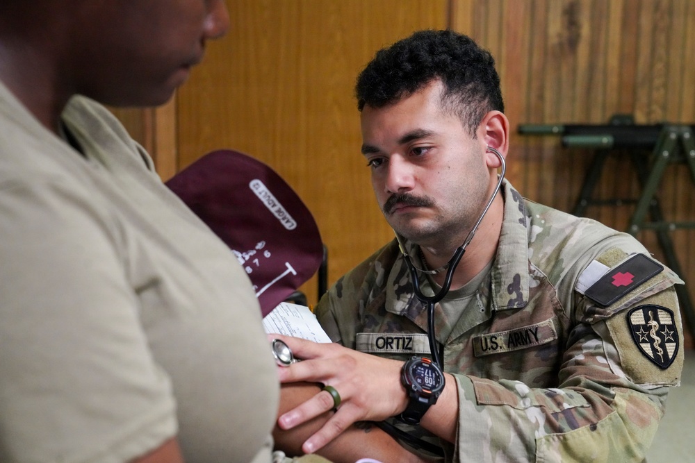A U.S. Army medic named Ortiz is performing a medical examination, likely taking a blood pressure reading, on another individual in what appears to be an indoor military setting. The medic is wearing a stethoscope and a tactical watch while concentrating on the patient's arm. A medical red cross patch and a unit patch are visible on his uniform sleeve.