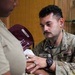 A U.S. Army medic named Ortiz is performing a medical examination, likely taking a blood pressure reading, on another individual in what appears to be an indoor military setting. The medic is wearing a stethoscope and a tactical watch while concentrating on the patient's arm. A medical red cross patch and a unit patch are visible on his uniform sleeve.