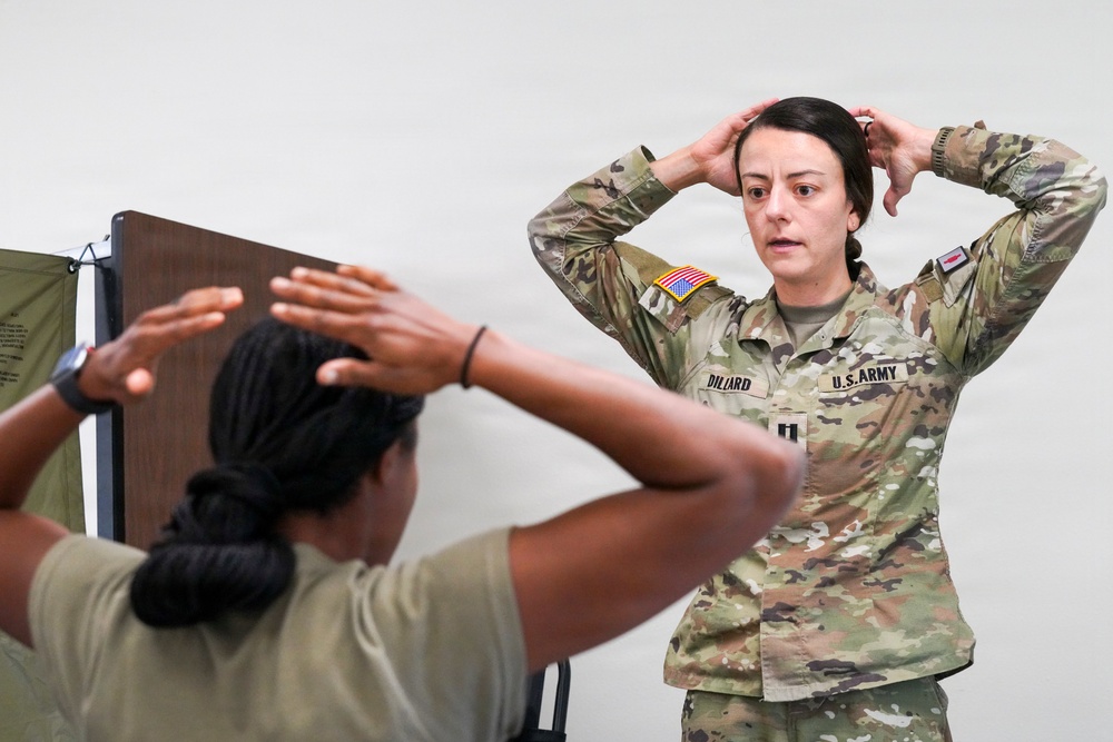 A U.S. Army soldier identified by name tape as 'Dillard' wearing OCP (Operational Camouflage Pattern) uniform with captain rank insignia is demonstrating a hands-on-head position to another soldier in a t-shirt who is mirroring the movement. The setting appears to be an indoor training or instruction environment. The interaction suggests a demonstration or physical assessment activity.