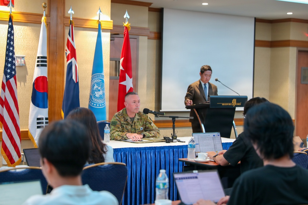 A formal military briefing or press conference is taking place in a conference room, likely at Dragon Hill Lodge in Seoul, South Korea. A civilian speaker addresses attendees from a podium while a military officer in camouflage uniform sits at a panel table. The presence of multiple national flags indicates a multinational military context.