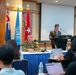 A formal military briefing or press conference is taking place in a conference room, likely at Dragon Hill Lodge in Seoul, South Korea. A civilian speaker addresses attendees from a podium while a military officer in camouflage uniform sits at a panel table. The presence of multiple national flags indicates a multinational military context.