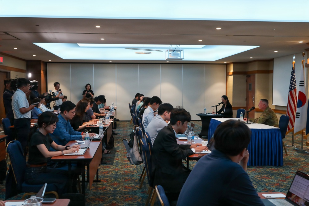 A formal press briefing or conference is taking place in a hotel-style conference room. A military officer in camouflage uniform and a civilian woman are seated at a head table facing an audience of journalists and attendees. The presence of both the American and South Korean flags suggests a U.S.-South Korea joint military or diplomatic event.