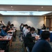 A formal press briefing or conference is taking place in a hotel-style conference room. A military officer in camouflage uniform and a civilian woman are seated at a head table facing an audience of journalists and attendees. The presence of both the American and South Korean flags suggests a U.S.-South Korea joint military or diplomatic event.