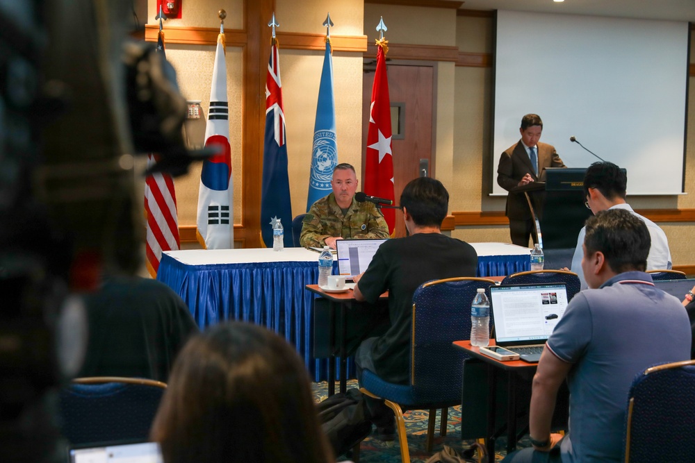 A military press briefing or conference is taking place in a formal meeting room. A uniformed military officer sits at a draped table facing journalists who are using laptops, while a civilian interpreter or moderator stands at a podium. Multiple national flags are displayed in the background, indicating a multinational or United Nations-related military context.
