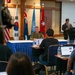 A military press briefing or conference is taking place in a formal meeting room. A uniformed military officer sits at a draped table facing journalists who are using laptops, while a civilian interpreter or moderator stands at a podium. Multiple national flags are displayed in the background, indicating a multinational or United Nations-related military context.