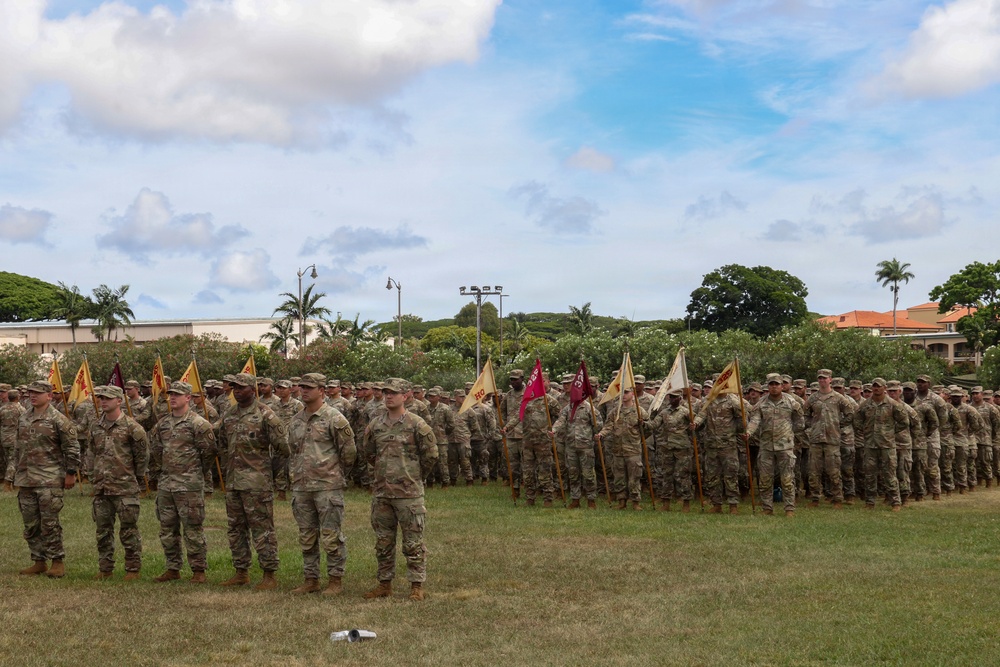 A large formation of U.S. Army soldiers in MultiCam/OCP camouflage uniforms are assembled on a grass parade field during a formal military ceremony. Multiple unit guidons (flags) in gold, red, and white are visible throughout the formation, indicating multiple units are represented. The tropical vegetation, palm trees, and architectural style in the background suggest this is located at a military installation in Hawaii, likely Schofield Barracks.