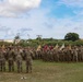 A large formation of U.S. Army soldiers in MultiCam/OCP camouflage uniforms are assembled on a grass parade field during a formal military ceremony. Multiple unit guidons (flags) in gold, red, and white are visible throughout the formation, indicating multiple units are represented. The tropical vegetation, palm trees, and architectural style in the background suggest this is located at a military installation in Hawaii, likely Schofield Barracks.