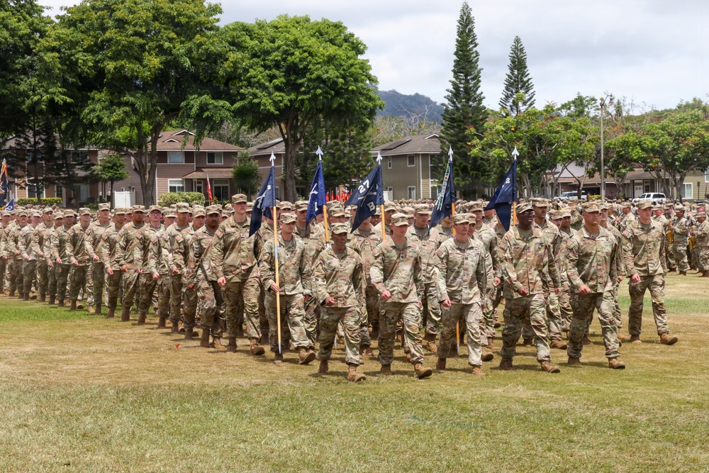 A large formation of U.S. Army soldiers in OCP (Operational Camouflage Pattern) uniforms marches across a grass field during what appears to be a formal military parade or ceremony. Multiple guidon bearers carry dark blue unit flags with gold finials at the front of the formation. The setting appears to be a military installation, likely in Hawaii based on the tropical vegetation and mountainous terrain visible in the background.