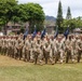 A large formation of U.S. Army soldiers in OCP (Operational Camouflage Pattern) uniforms marches across a grass field during what appears to be a formal military parade or ceremony. Multiple guidon bearers carry dark blue unit flags with gold finials at the front of the formation. The setting appears to be a military installation, likely in Hawaii based on the tropical vegetation and mountainous terrain visible in the background.