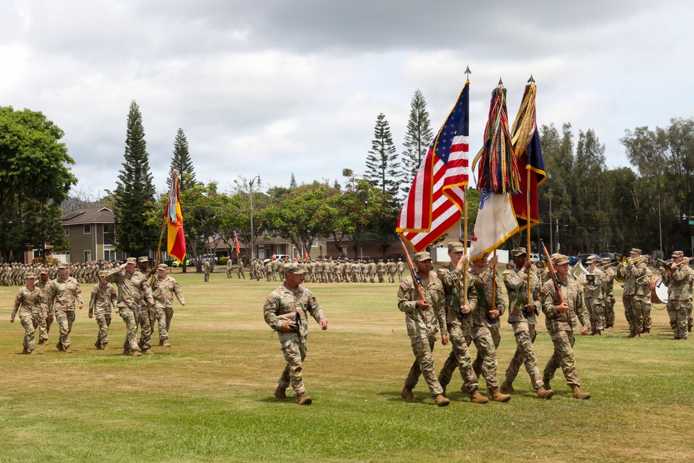 A U.S. Army military parade or ceremony is taking place on a large grass field, likely at a base in Hawaii based on the tropical vegetation including Norfolk Island pines. A color guard leads the formation carrying multiple flags including the American flag and unit guidons. Large formations of soldiers in Army Combat Uniforms (ACU/OCP) are visible in the background, along with a military band on the right.