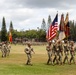 A U.S. Army military parade or ceremony is taking place on a large grass field, likely at a base in Hawaii based on the tropical vegetation including Norfolk Island pines. A color guard leads the formation carrying multiple flags including the American flag and unit guidons. Large formations of soldiers in Army Combat Uniforms (ACU/OCP) are visible in the background, along with a military band on the right.