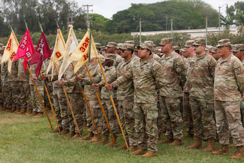A formal U.S. Army military formation is taking place on a grass field, with soldiers standing at attention in multicam/OCP camouflage uniforms. Multiple guidon bearers are holding unit flags displaying the number '325' along with company designations including 'HQ', 'B', and 'D'. The setting appears to be a military installation, likely in a tropical or subtropical region based on the vegetation visible in the background.