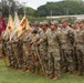 A formal U.S. Army military formation is taking place on a grass field, with soldiers standing at attention in multicam/OCP camouflage uniforms. Multiple guidon bearers are holding unit flags displaying the number '325' along with company designations including 'HQ', 'B', and 'D'. The setting appears to be a military installation, likely in a tropical or subtropical region based on the vegetation visible in the background.