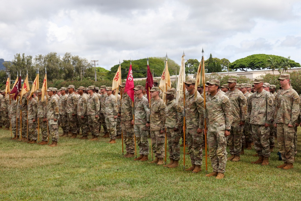 A formal U.S. Army military formation ceremony is taking place on a grass field, likely at a base in Hawaii based on the tropical vegetation and terrain visible in the background. Soldiers in Operational Camouflage Pattern (OCP) uniforms are standing at attention in organized ranks, with guidon bearers holding unit flags (guidons) in red, maroon, and tan/yellow colors. The event appears to be a change of command, unit activation, or similar official military ceremony.