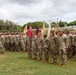 A formal U.S. Army military formation ceremony is taking place on a grass field, likely at a base in Hawaii based on the tropical vegetation and terrain visible in the background. Soldiers in Operational Camouflage Pattern (OCP) uniforms are standing at attention in organized ranks, with guidon bearers holding unit flags (guidons) in red, maroon, and tan/yellow colors. The event appears to be a change of command, unit activation, or similar official military ceremony.