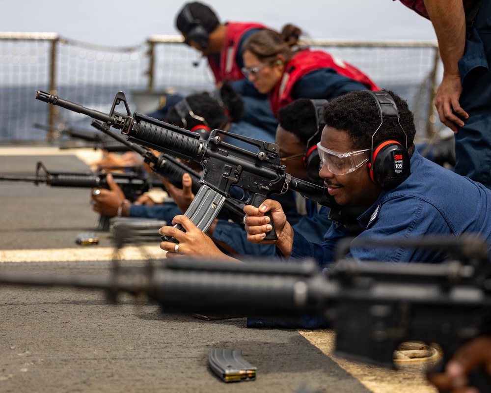 Military personnel are conducting a weapons qualification or firearms training exercise on what appears to be the deck of a naval vessel. Multiple individuals in blue Navy coveralls are in the prone position aiming M16/M4-style rifles downrange. Safety equipment including hearing protection and eye protection is being worn by the participants.