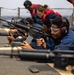 Military personnel are conducting a weapons qualification or firearms training exercise on what appears to be the deck of a naval vessel. Multiple individuals in blue Navy coveralls are in the prone position aiming M16/M4-style rifles downrange. Safety equipment including hearing protection and eye protection is being worn by the participants.