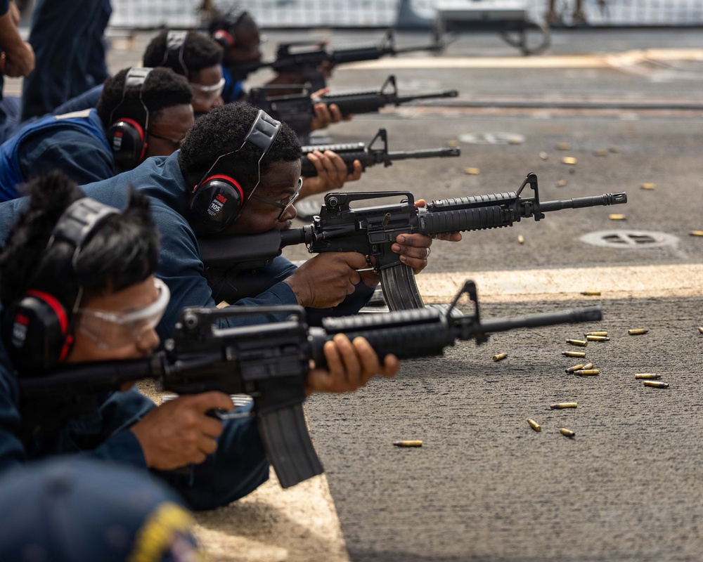 A group of military personnel in blue uniforms are conducting a rifle qualification or training exercise on what appears to be the deck of a naval vessel. Multiple shooters are in a prone or low-position firing stance, aiming M4/M16-style carbines downrange. Spent brass casings are visible on the deck surface, indicating active firing.
