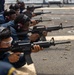 A group of military personnel in blue uniforms are conducting a rifle qualification or training exercise on what appears to be the deck of a naval vessel. Multiple shooters are in a prone or low-position firing stance, aiming M4/M16-style carbines downrange. Spent brass casings are visible on the deck surface, indicating active firing.