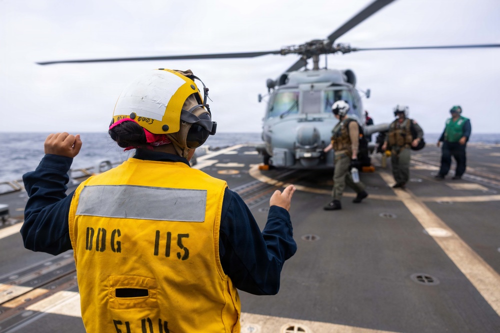 A naval flight deck crew member wearing a yellow safety vest marked 'DDG 115' and a white helmet is directing helicopter operations on a ship's flight deck. A military helicopter, likely an MH-60 Seahawk, is visible in the background with crew members and pilots nearby. The scene takes place at sea under overcast skies.