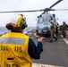A naval flight deck crew member wearing a yellow safety vest marked 'DDG 115' and a white helmet is directing helicopter operations on a ship's flight deck. A military helicopter, likely an MH-60 Seahawk, is visible in the background with crew members and pilots nearby. The scene takes place at sea under overcast skies.