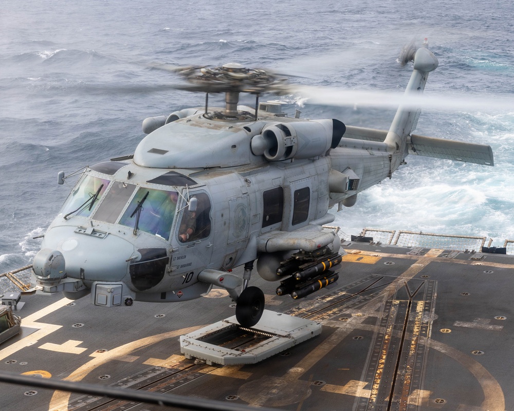 A U.S. Navy MH-60R Seahawk helicopter is hovering over the flight deck of a naval vessel at sea. The helicopter is armed with what appear to be AGM-114 Hellfire missiles on its starboard weapons pylon. The rotor blades are in motion, and the ocean is visible in the background with white wake from the ship.